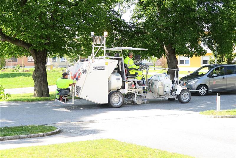 Macchina traccialinee con due operatori in giubbotto ad alta visibilità, uno alla guida e uno seduto a bordo, su una strada di quartiere alberata